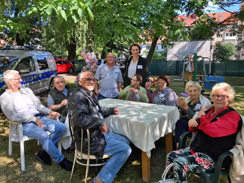 seniorzy siedzą w ogrodzie przy stoliku podczas imprezy, na zdjeciu z panią prezydentką Moniką Chabior